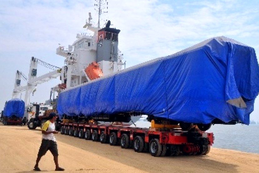 Trailer multi-axle membawa kargo panjang yang tertutup terpal biru di pelabuhan dengan kapal di latar belakang